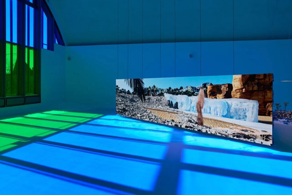 10 Exhibitions Not to Miss During Mexico City Art Week 2026 A wide interior gallery space is bathed in blue and green light from large colored windows on the left, with a long horizontal video screen on the floor showing a desert-like landscape with rocks, ice formations, and a human figure, casting reflections across the polished floor.