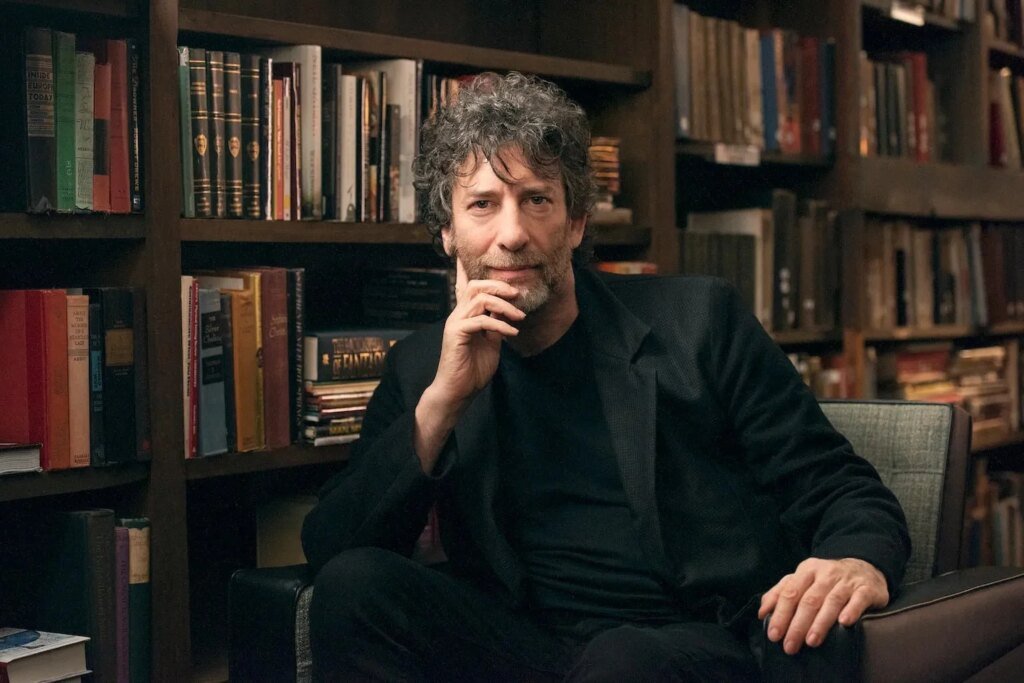 Neil Gaiman is sitting on a chair with rows of books behind him