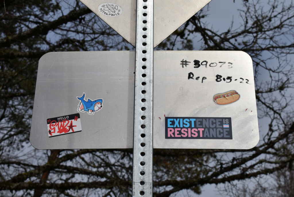 The back of a street sign is covered in stickers. On the bottom left corner, one stands out saying "Existence is Resistance” with the trans pride flag and its colors used in the message. The phrase has been used across many movements, but with this particular sticker is a nod to the attacks on trans rights across the U.S. (Emma Kahl/Daily Emerald)